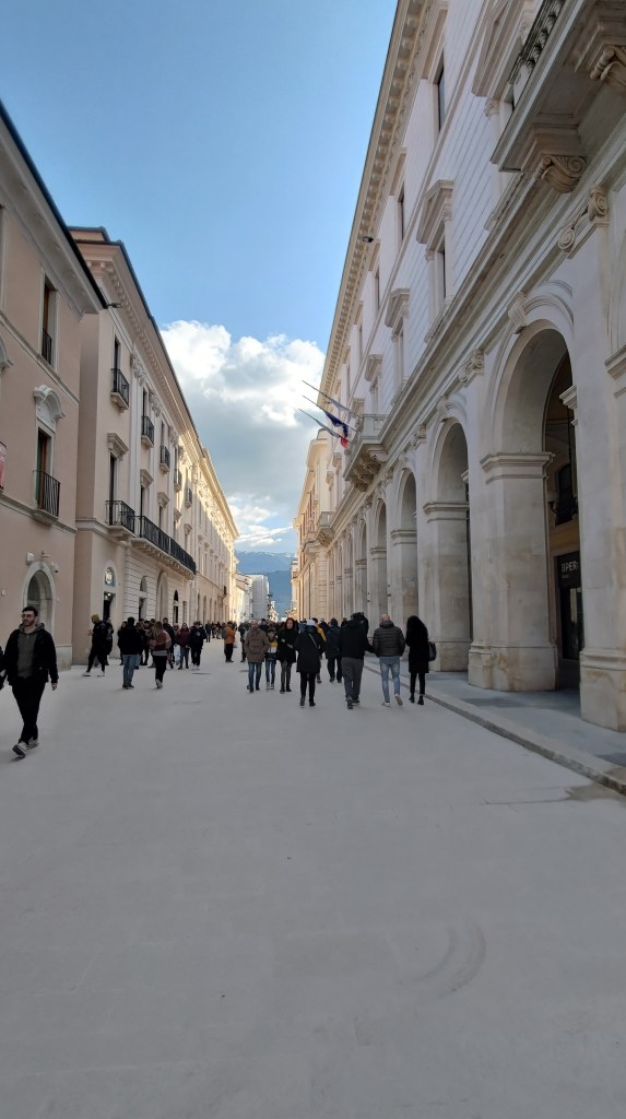 Il centro storico dell'Aquila, con persone che passeggiano lungo una strada pedonale fiancheggiata da edifici storici, sotto un cielo parzialmente nuvoloso.
